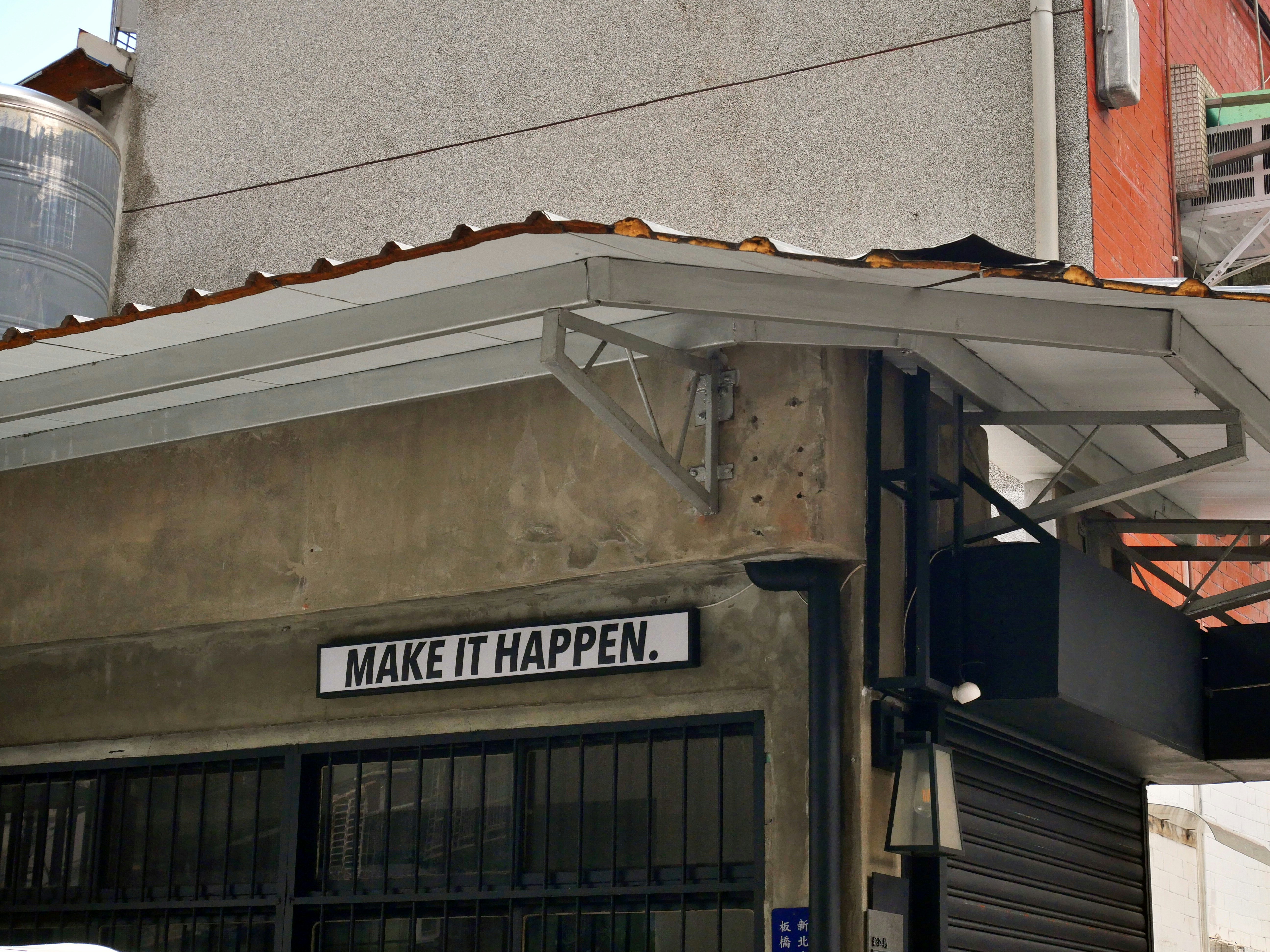 A sign high up on a wall on a street corner reads in English "Make it Happen".